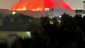 A pyramid-shaped structure with a red glow, surrounded by trees and buildings. - Regent Pyramids View (Giza)
