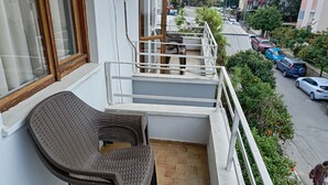 Un balcón con una silla de mimbre, una mesita y vistas a coches aparcados y zonas verdes en la calle.