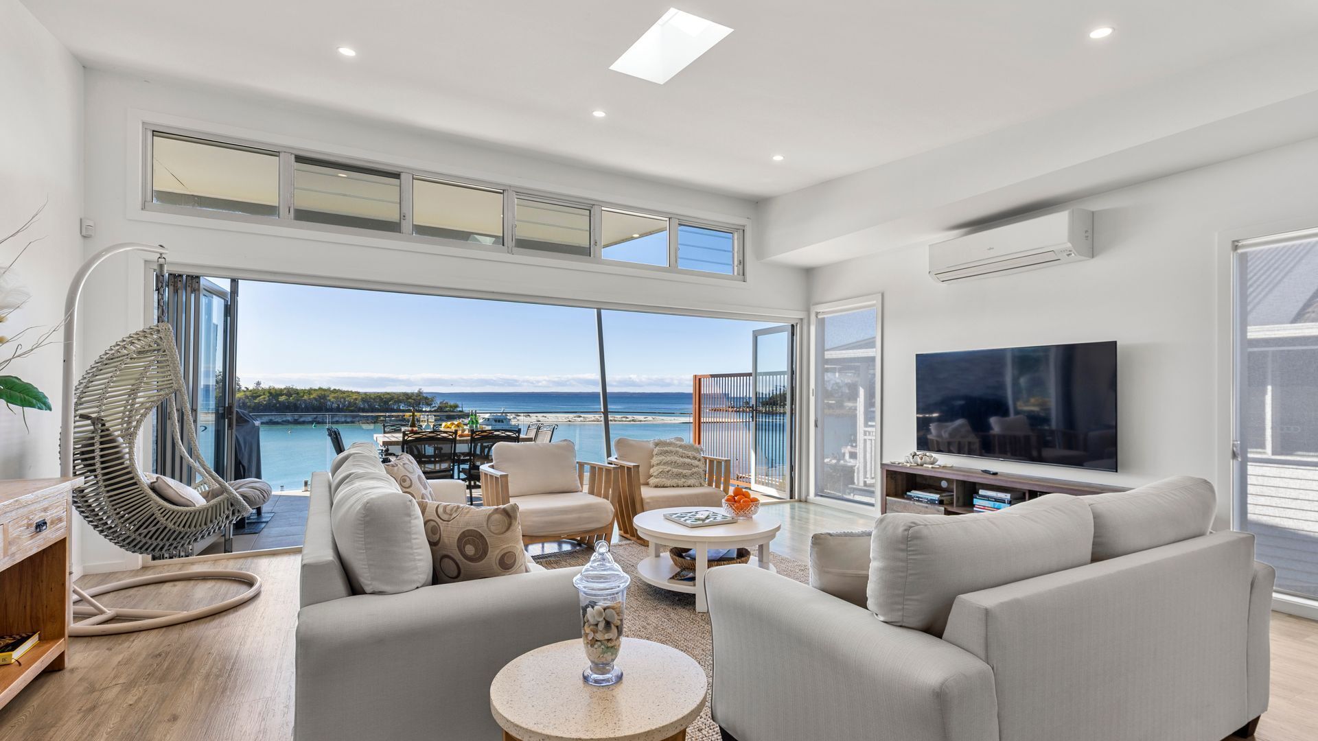 A modern living room with a large TV, comfortable seating, and a view of the sea.
