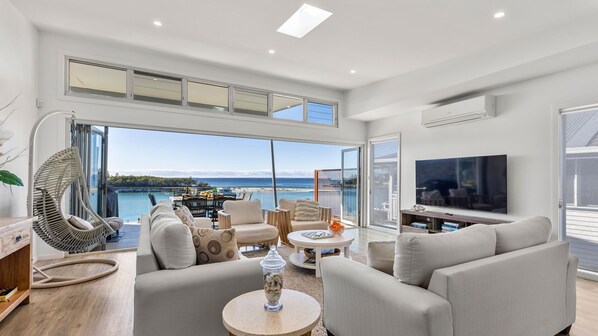 A modern living room with a large TV, comfortable seating, and a view of the sea.
