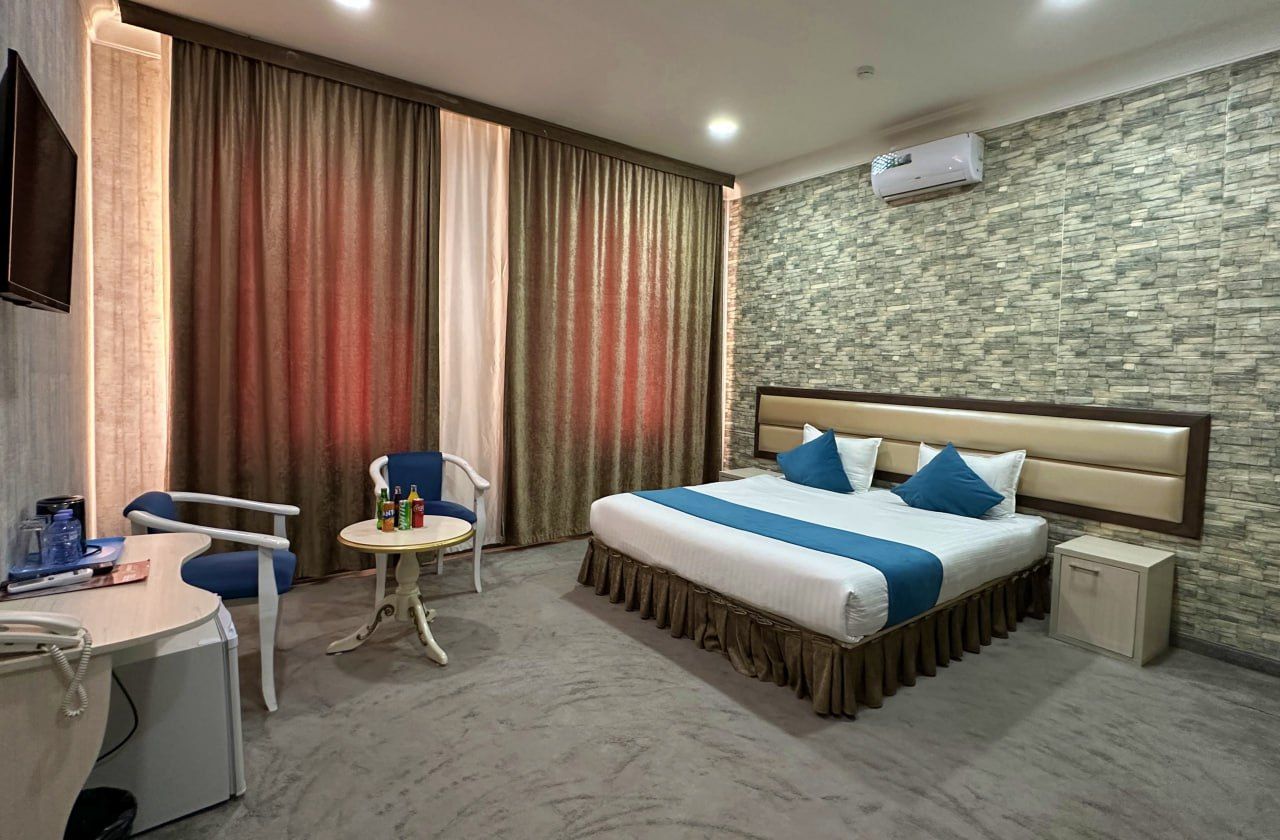 A hotel room with a bed, a desk, a chair, a small table, and a wall-mounted air conditioner.