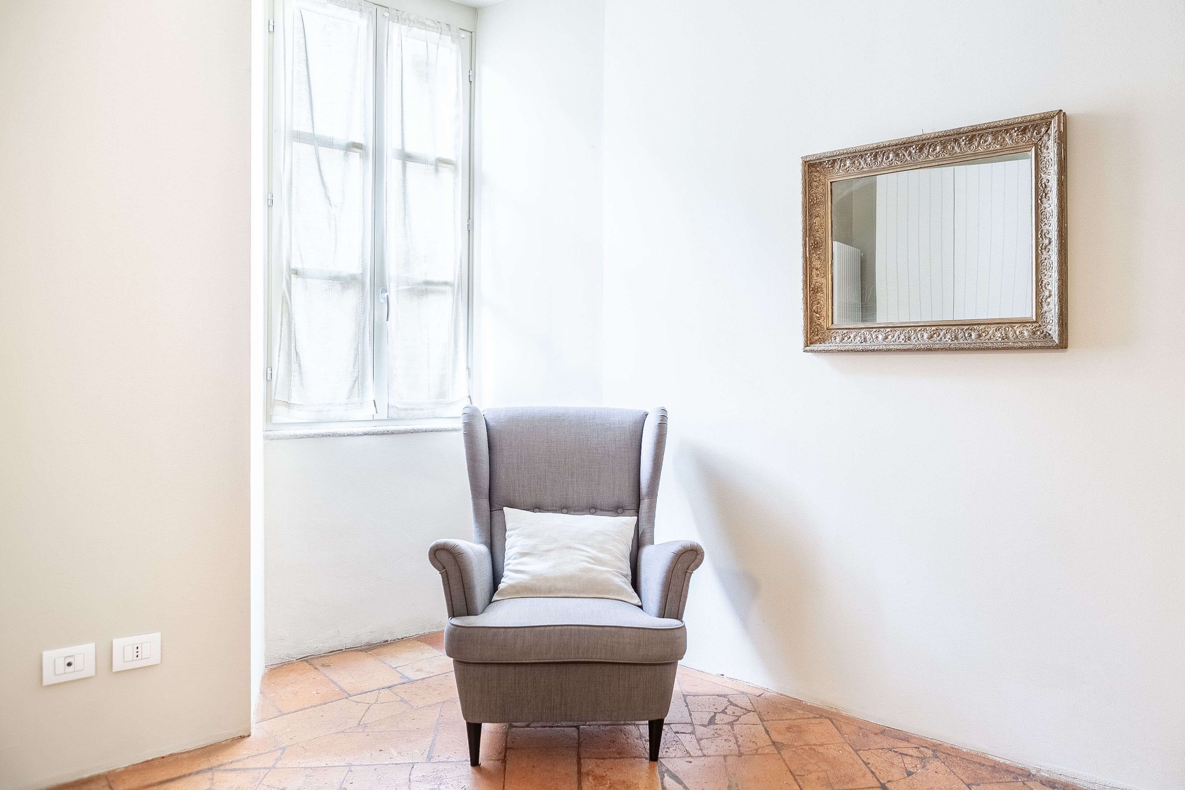 A room with a grey armchair, a mirror with an ornate frame, and a window with curtains.