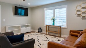 A living room with a ceiling fan, a flat-screen TV, a white entertainment unit, a brown leather sofa, a patterned rug, and a window with blinds.