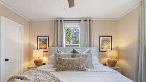 A bedroom with a bed, two bedside lamps, a window with curtains, and framed pictures on the wall. - Swamp Rabbit | Furman | Bikes | Pass | Fenced | EV (Greenville)