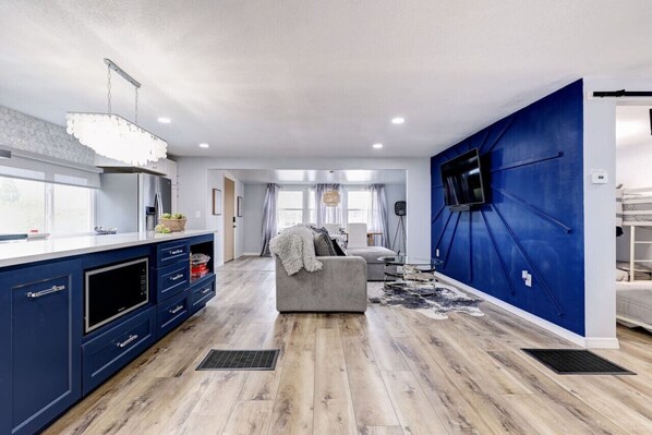 Eine moderne Küche mit integriertem Mikrowellenherd, einer großen Kochinsel mit Spüle sowie einem Essbereich mit grauem Sofa und an einer blauen Akzentwand montiertem Fernseher.