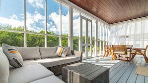 A covered outdoor seating area with a sofa, wicker ottoman, and wooden chairs.