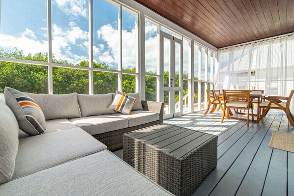 A covered outdoor seating area with a sofa, wicker ottoman, and wooden chairs.