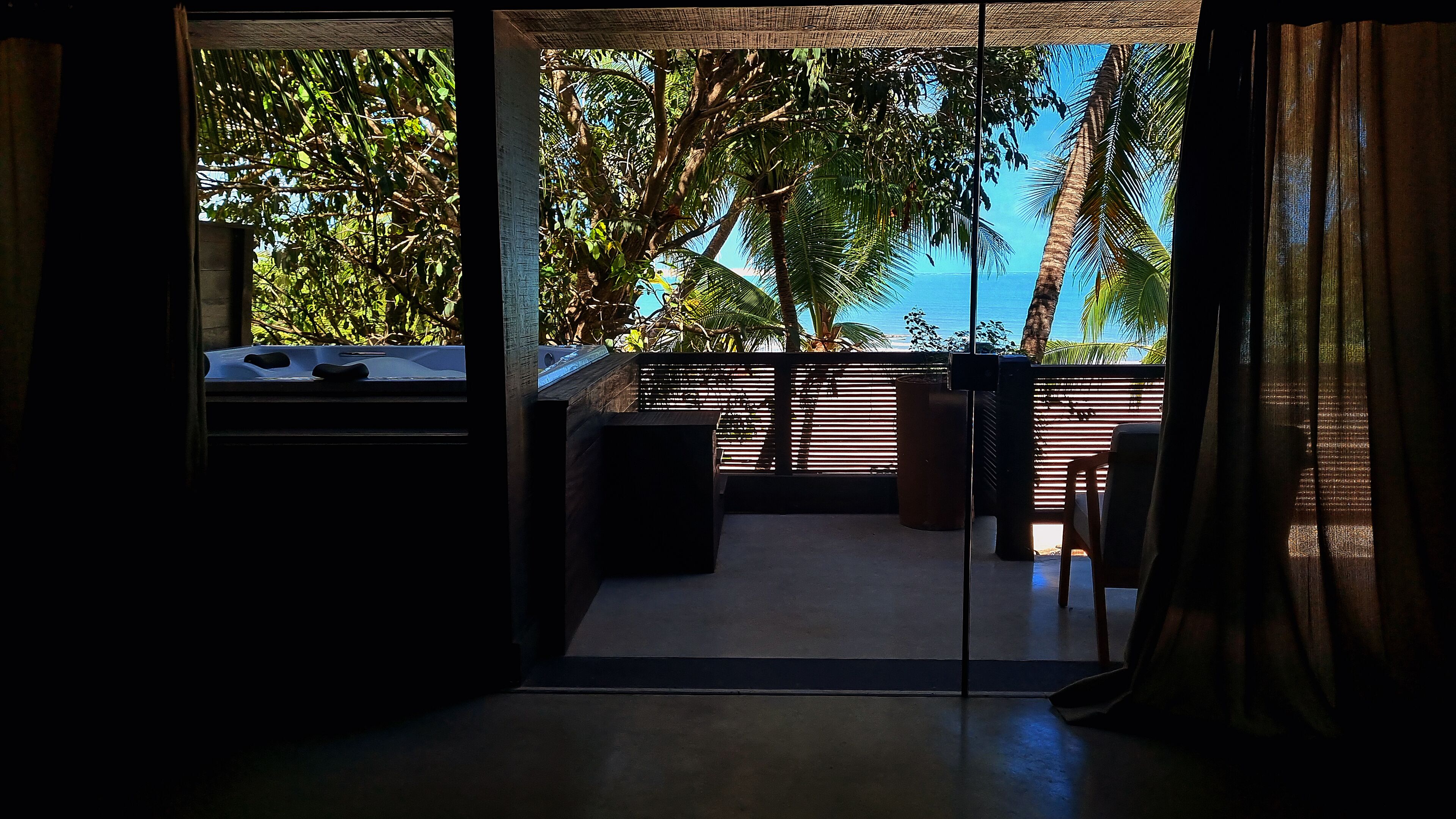 A room with a view of palm trees and the sea. There is a small seating area with a bench and a table.