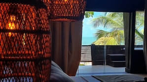 A room with a bed, two wicker lamps, a window with a view of palm trees and the sea, and a curtain.