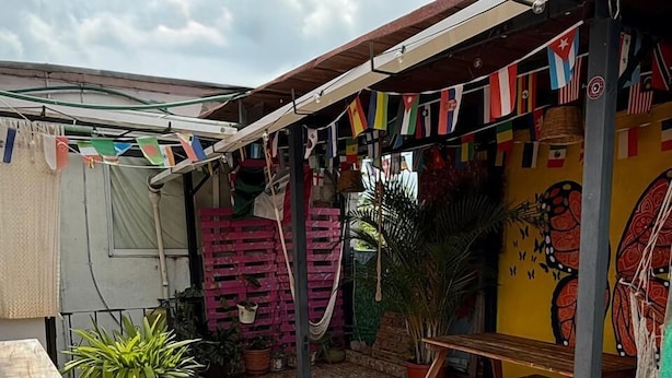 Un patio con suelo de baldosas, mobiliario de madera, plantas en macetas y una pared adornada con banderas de colores.