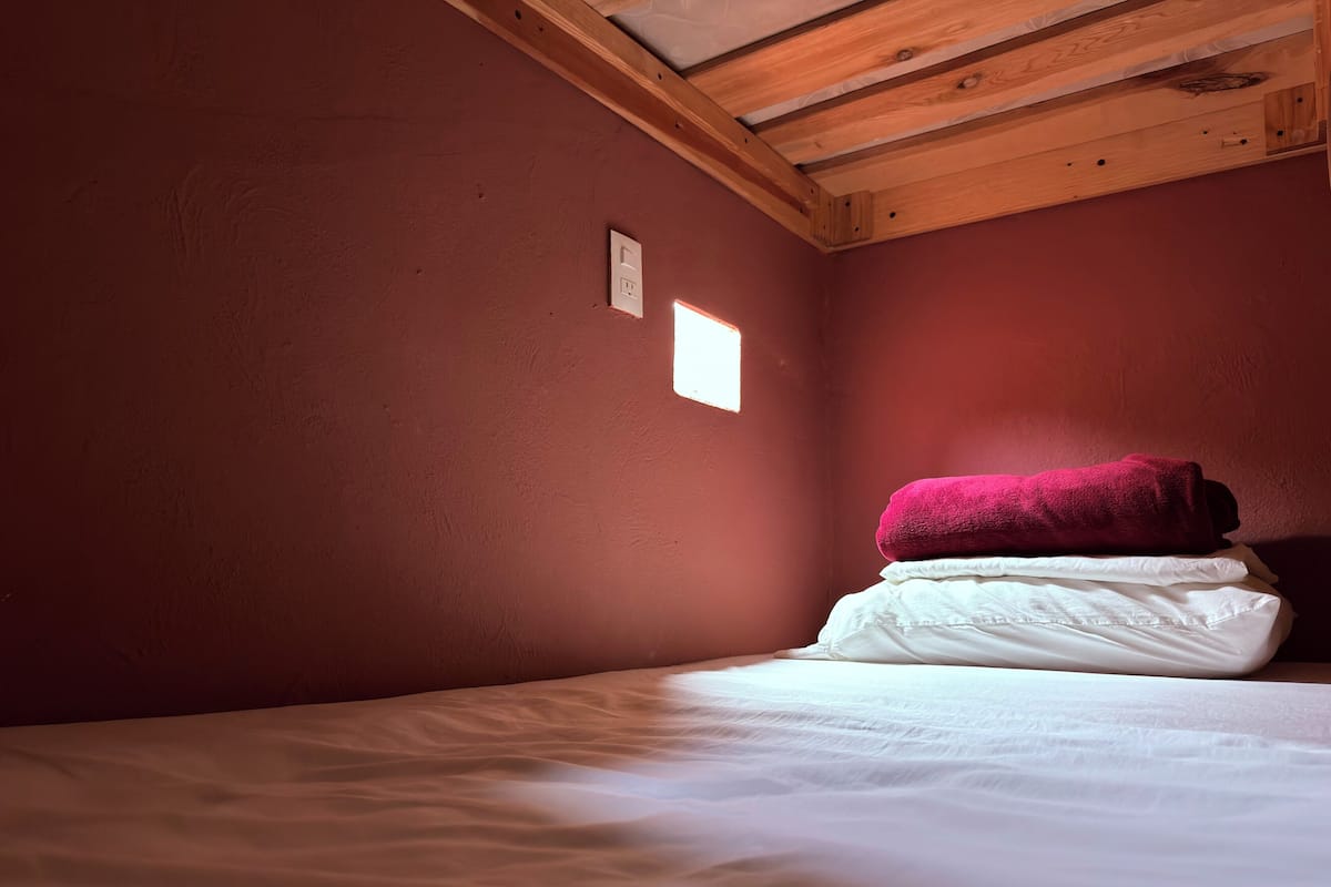 A room with a single bed, a wooden ceiling, and a small window.