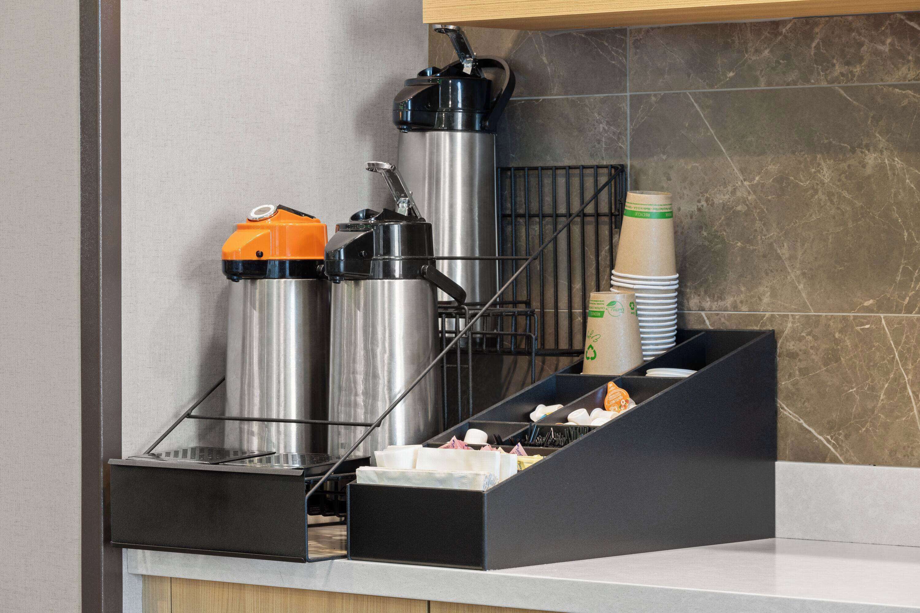 Un’area self-service per bevande con erogatori in acciaio inossidabile e una macchina per il caffè, una pila di bicchieri usa e getta e un vassoio con condimenti.