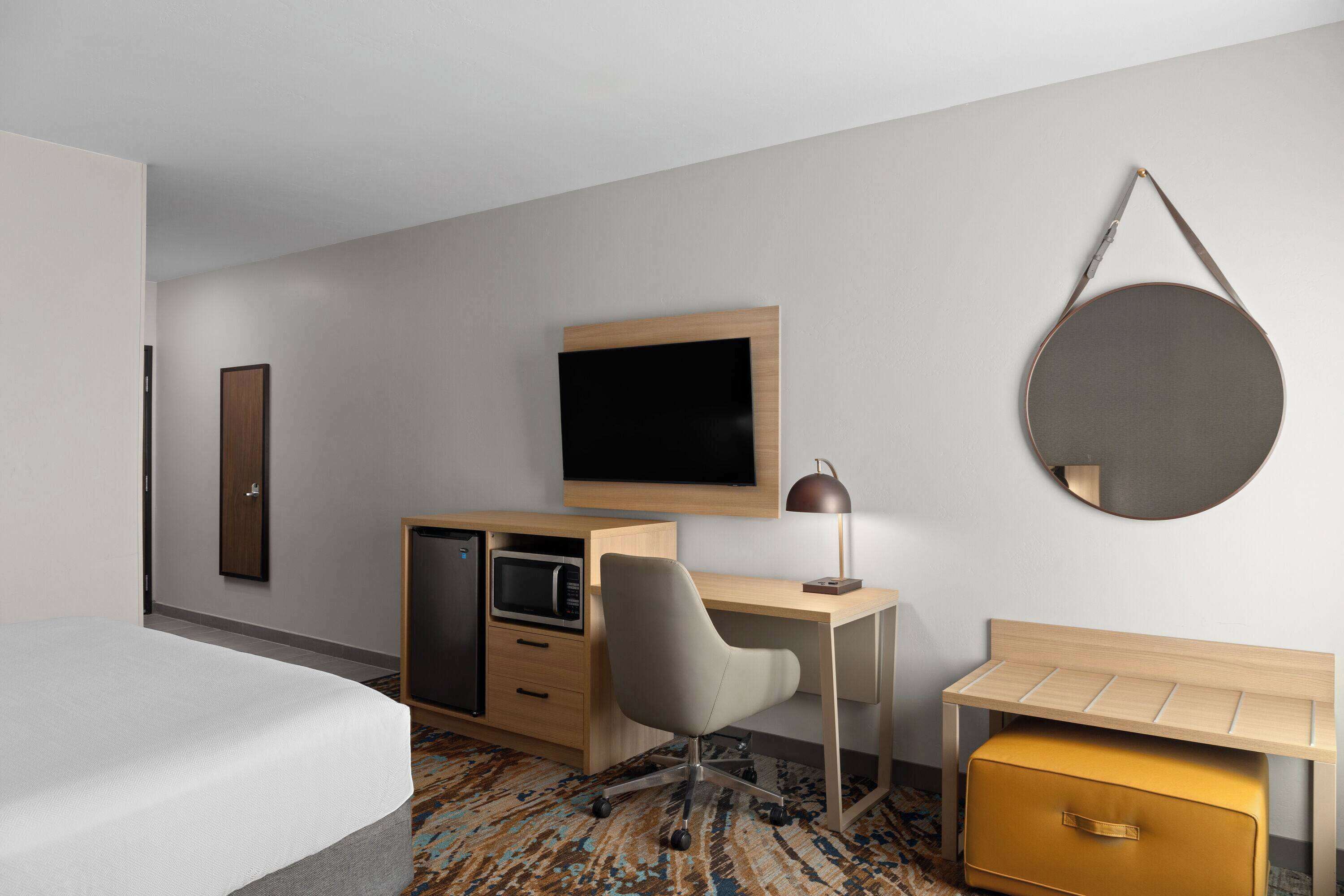 Una habitación de hotel con una cama, un escritorio con silla, un televisor, un microondas, una mesita y un espejo redondo en la pared.