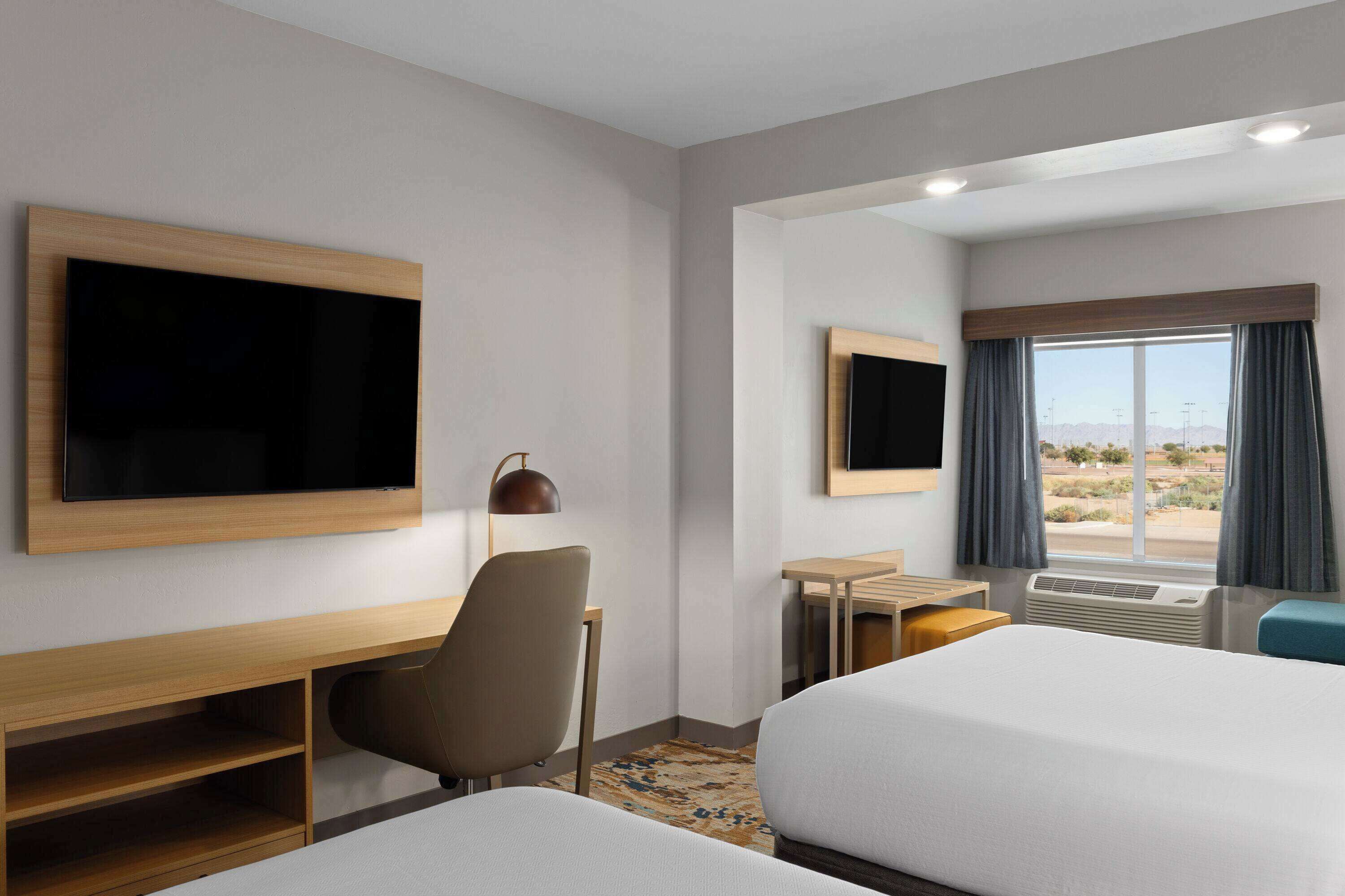 A hotel room with two beds, a desk, a chair, a TV mounted on the wall, and a window with curtains.