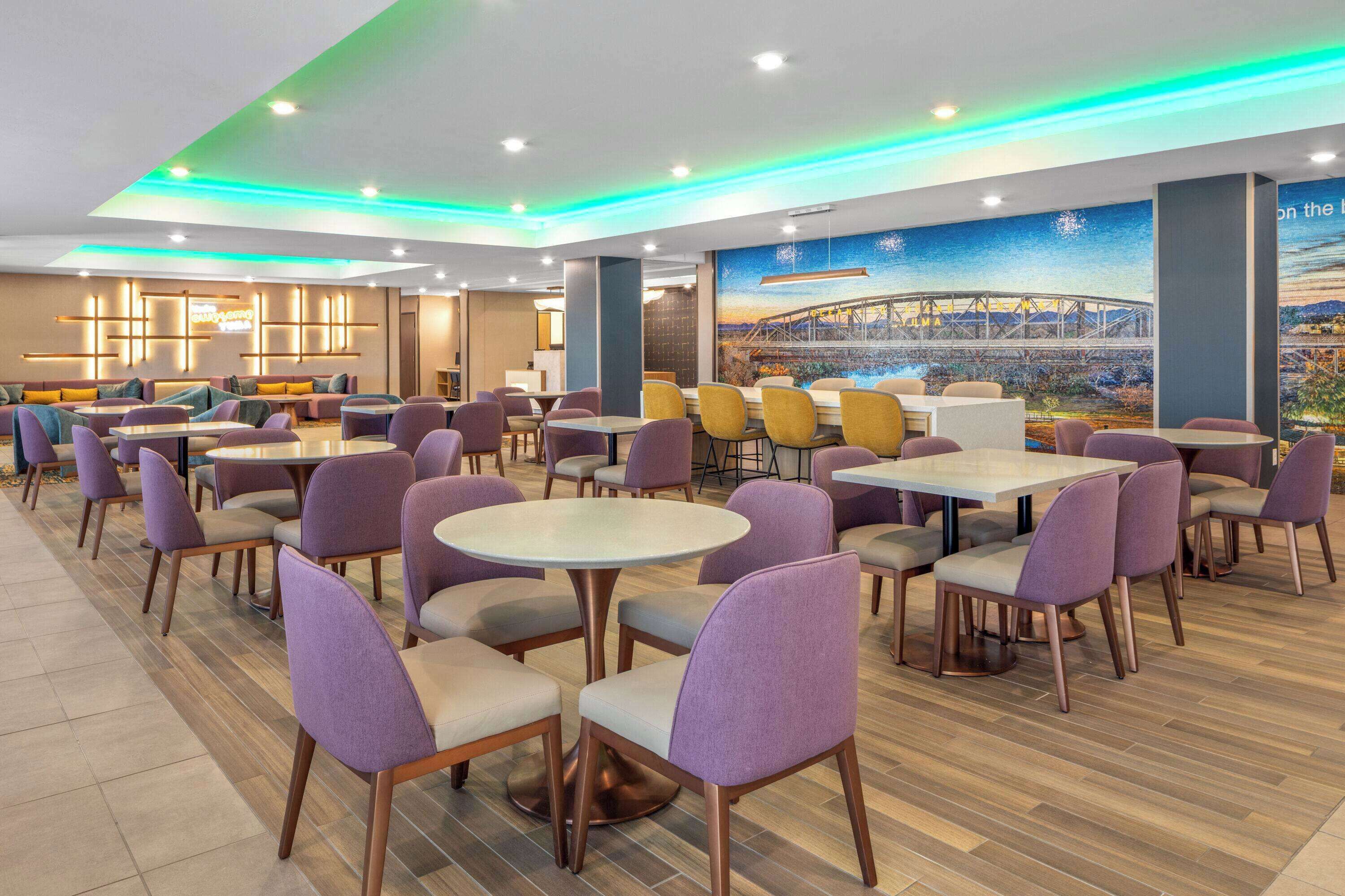 A modern dining area with round tables and purple chairs, featuring a mural of a bridge and cityscape on the wall.