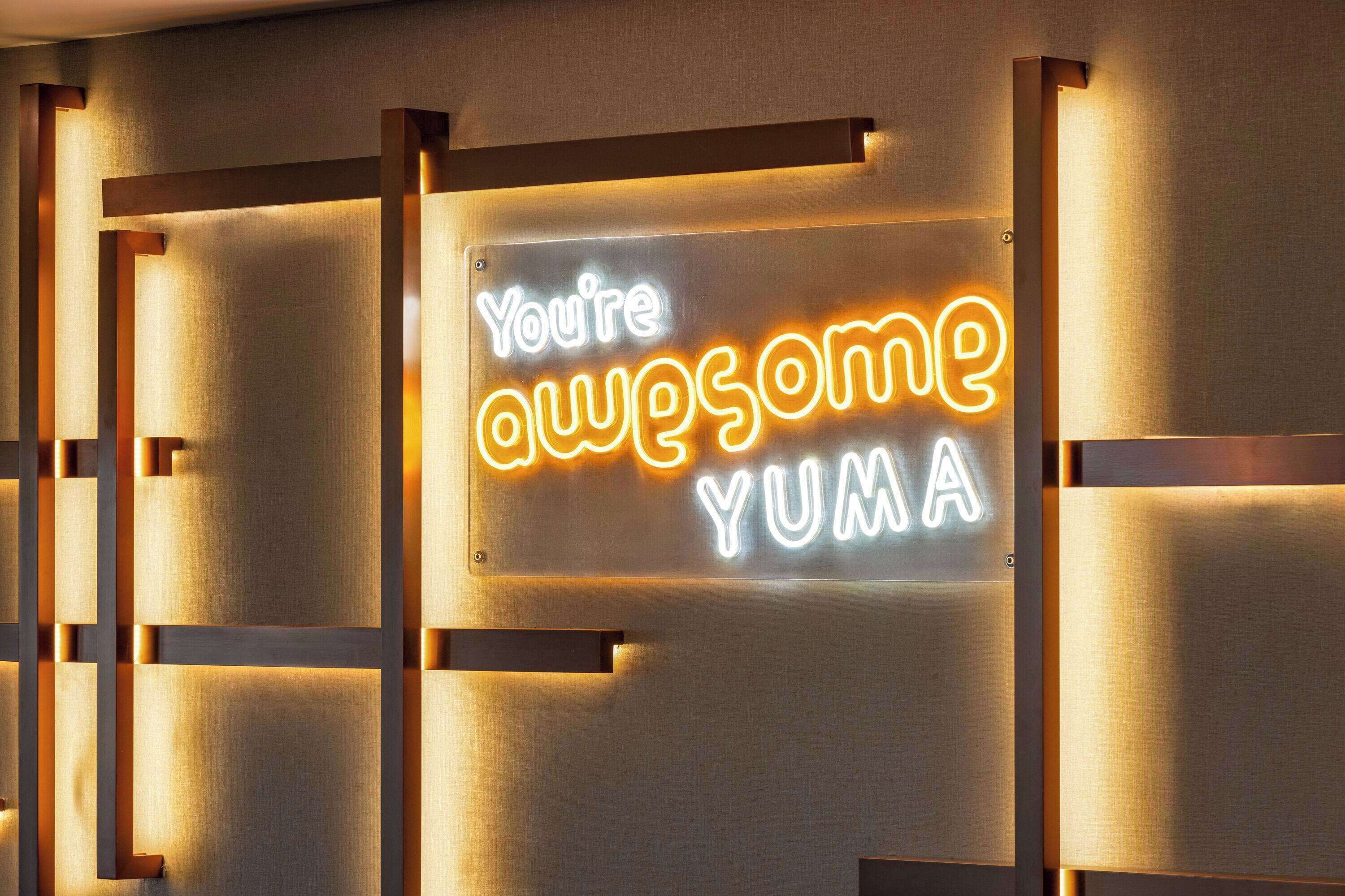 A neon sign with the text "You're awesome YUMA" on a wall with a geometric pattern.