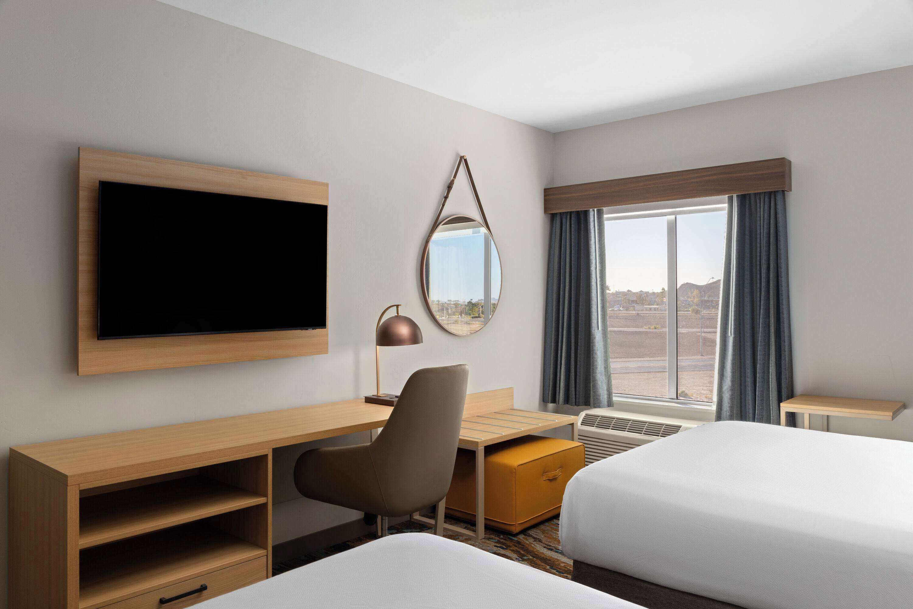 A hotel room with a bed, a desk with a chair, a TV, a mirror, and a window with curtains.