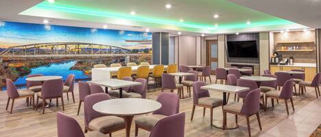 A hotel dining area with round tables and purple chairs, a large TV screen, and a mural of a bridge over water.