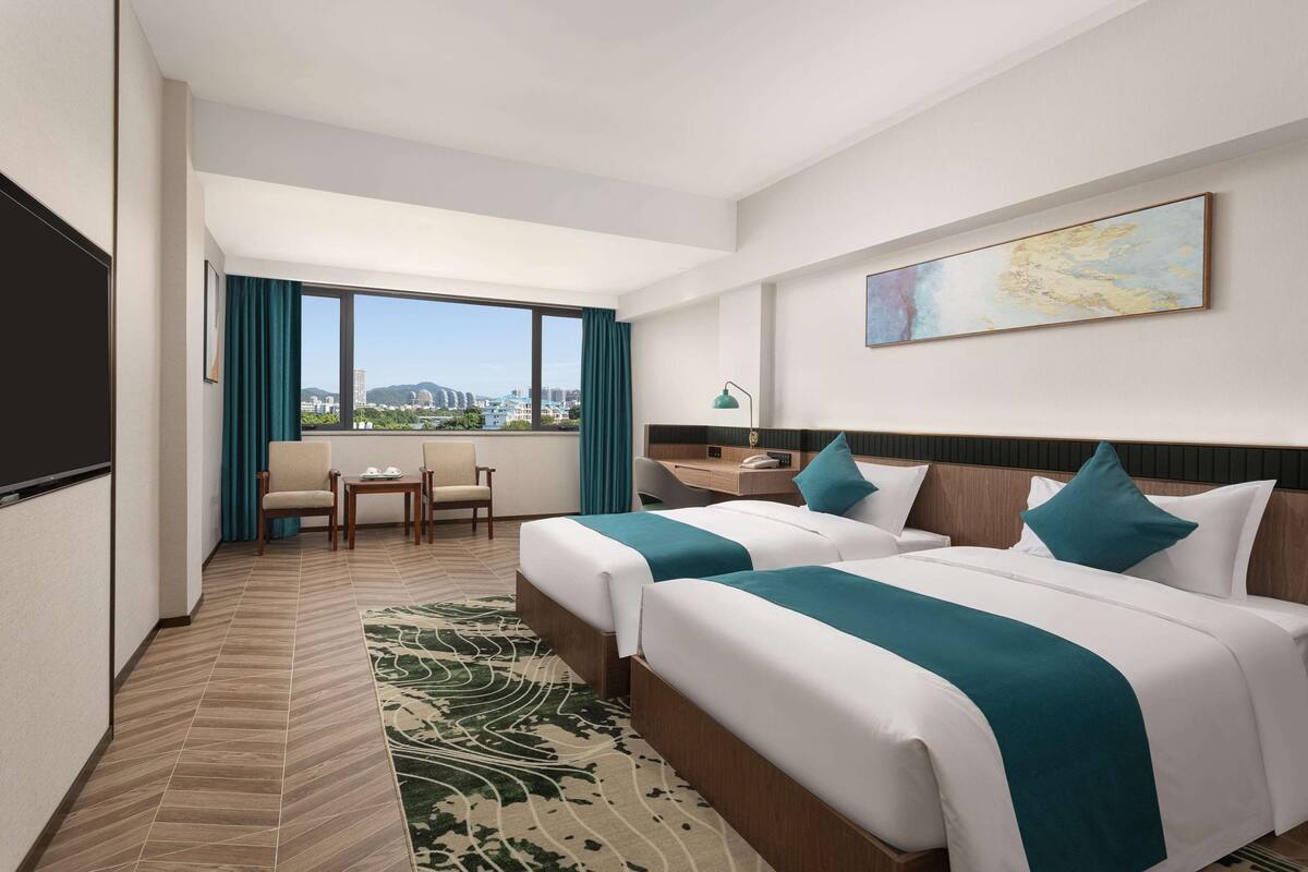 Une chambre d’hôtel avec deux lits, un bureau, une chaise, une grande fenêtre donnant sur un beau paysage et un tableau au mur.