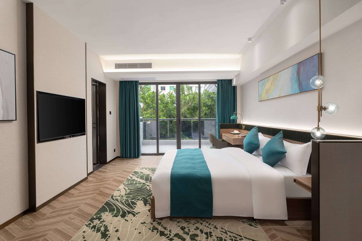 Une chambre d’hôtel moderne dotée d’un grand lit, d’un bureau, d’une télévision à écran plat et d’un balcon offrant une vue sur la verdure.
