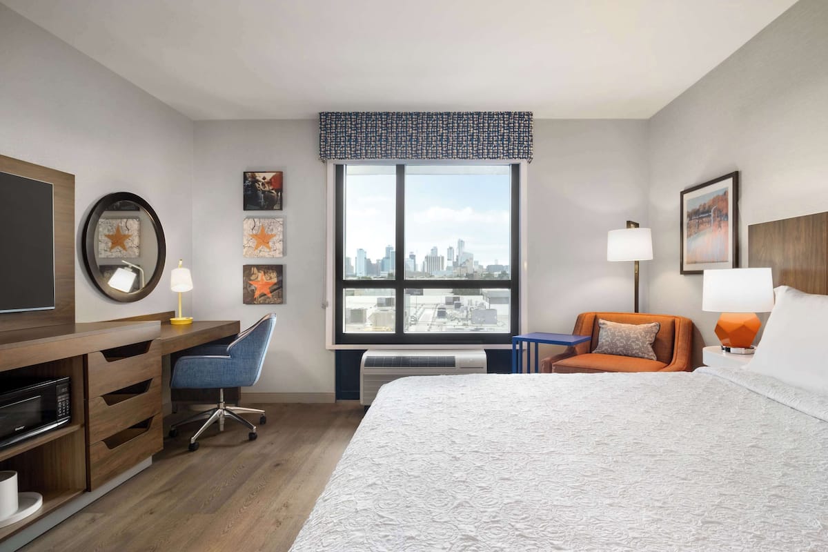 A hotel room with a large bed, a desk with a chair, a television, and a window with a city view.