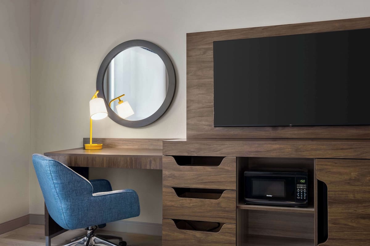 A modern hotel room with a wooden desk, a blue chair, a round mirror, a desk lamp, a flat-screen TV, and a built-in microwave.
