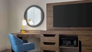 Una habitación de hotel moderna con un escritorio de madera, una silla azul, un espejo redondo, una lámpara de escritorio, un televisor de pantalla plana y un microondas integrado.