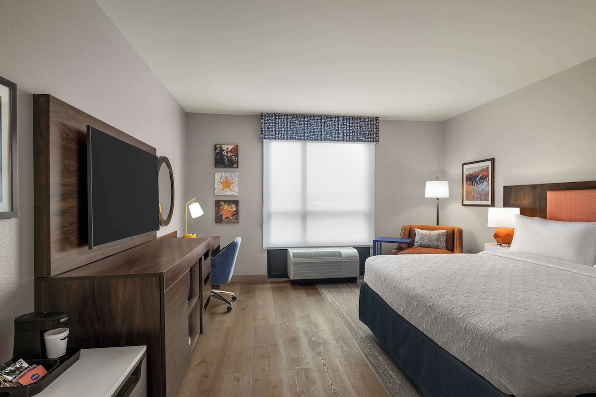 A hotel room with a bed, desk, chair, TV, air conditioning unit, and artwork on the walls.