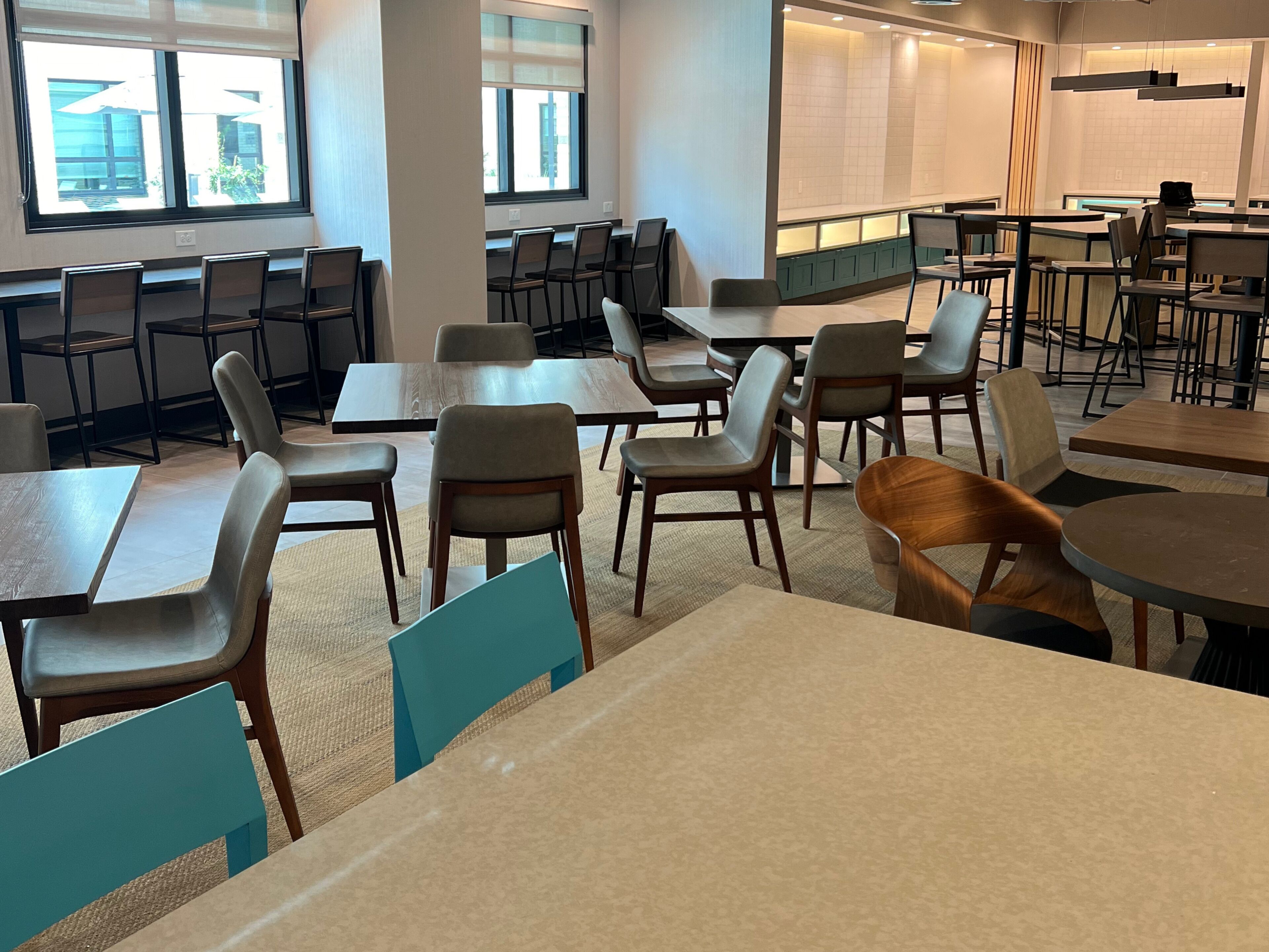 A spacious dining area with multiple tables and chairs, large windows providing natural light, and a modern kitchen area in the background.
