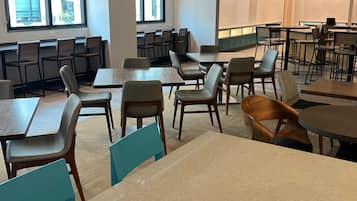 A spacious dining area with multiple tables and chairs, large windows providing natural light, and a modern kitchen area in the background.