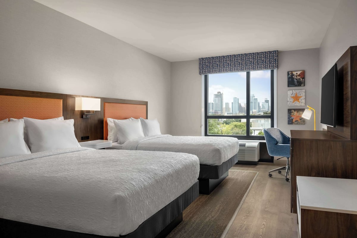 A hotel room with two beds, a desk, a chair, a TV, and a large window with a city view.