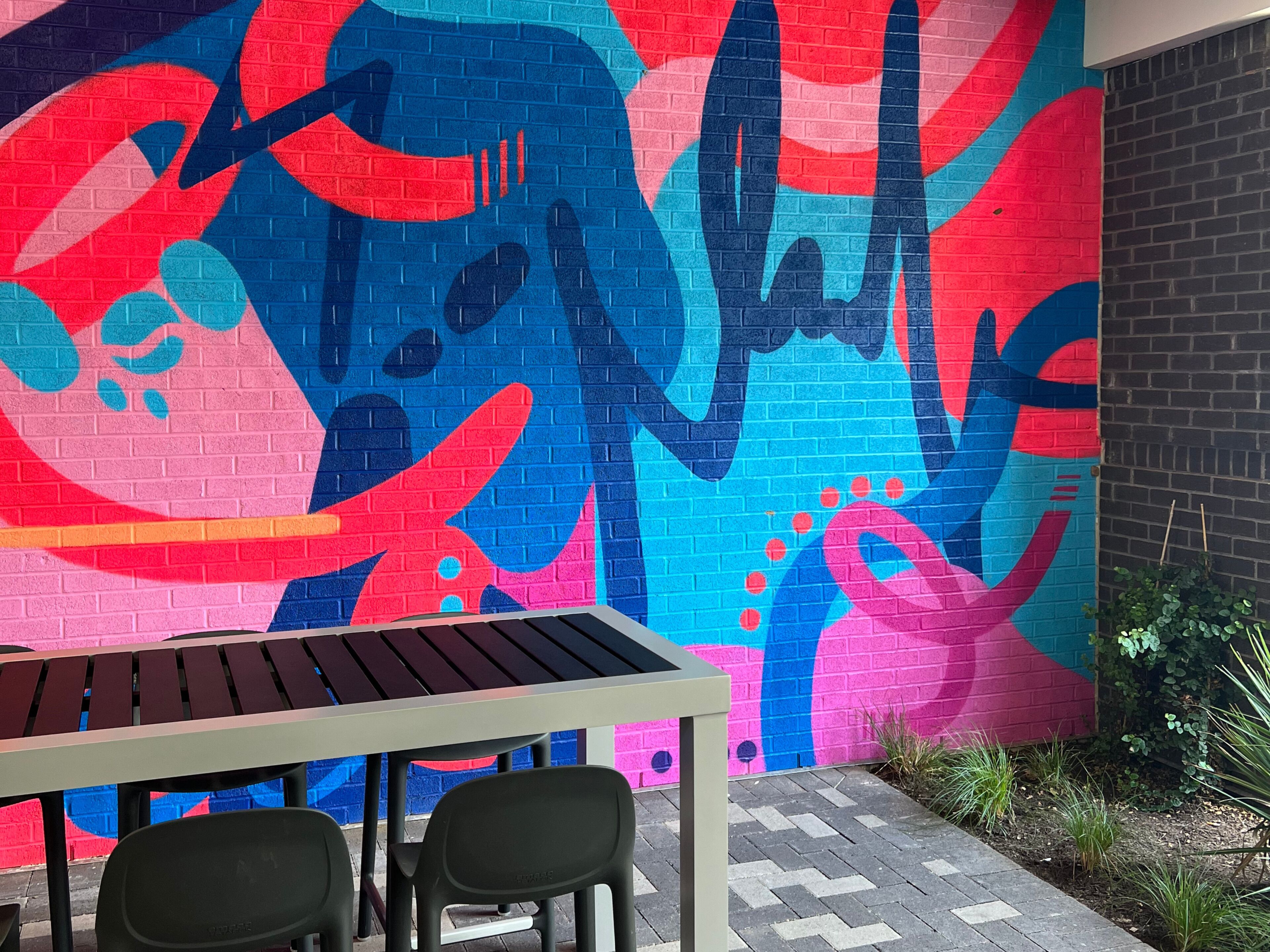 A dining area with a table and chairs, a colorful mural on the wall, and a brick wall to the right.