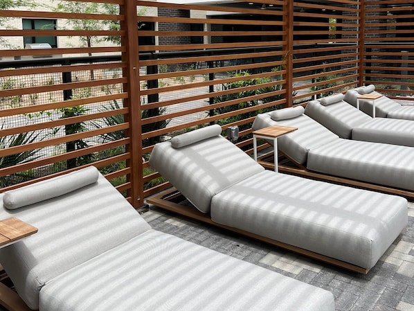 A rooftop lounge area with multiple grey cushioned lounge chairs and wooden tables, surrounded by a wooden lattice fence.