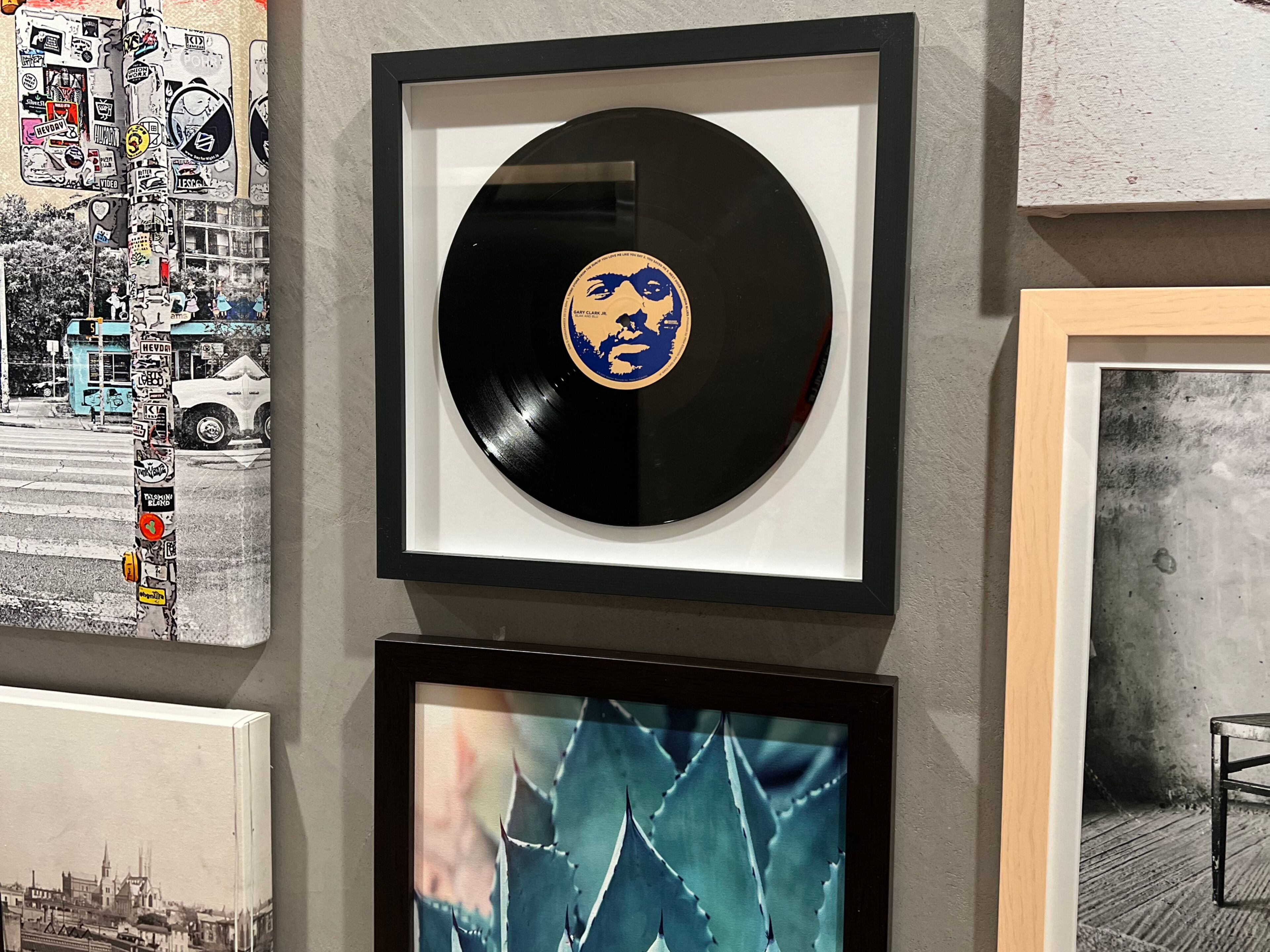 Eine gerahmte Vinylschallplatte mit blau-gelbem Design, ein gerahmtes Stadtbild und ein gerahmtes Pflanzenbild.