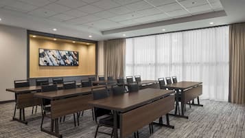 Une salle de conférence équipée de plusieurs tables et chaises, d’une grande fenêtre avec des rideaux et d’un écran fixé au mur.