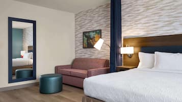Quarto de hotel com cama, sofá, puf redondo, espelho, quadro na parede e mesa de cabeceira com abajur.