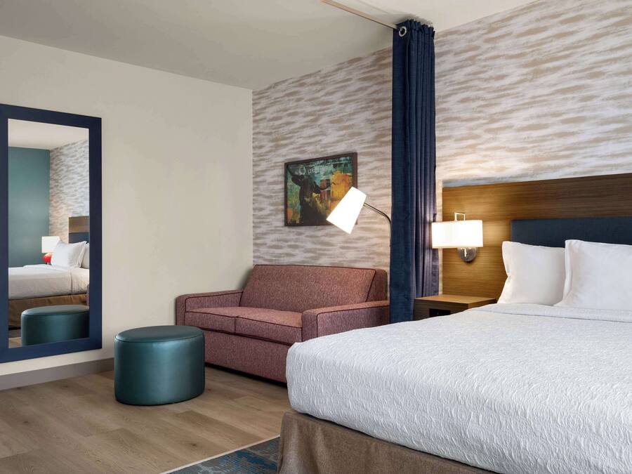Quarto de hotel com cama, sofá, puf redondo, espelho, quadro na parede e mesa de cabeceira com abajur.