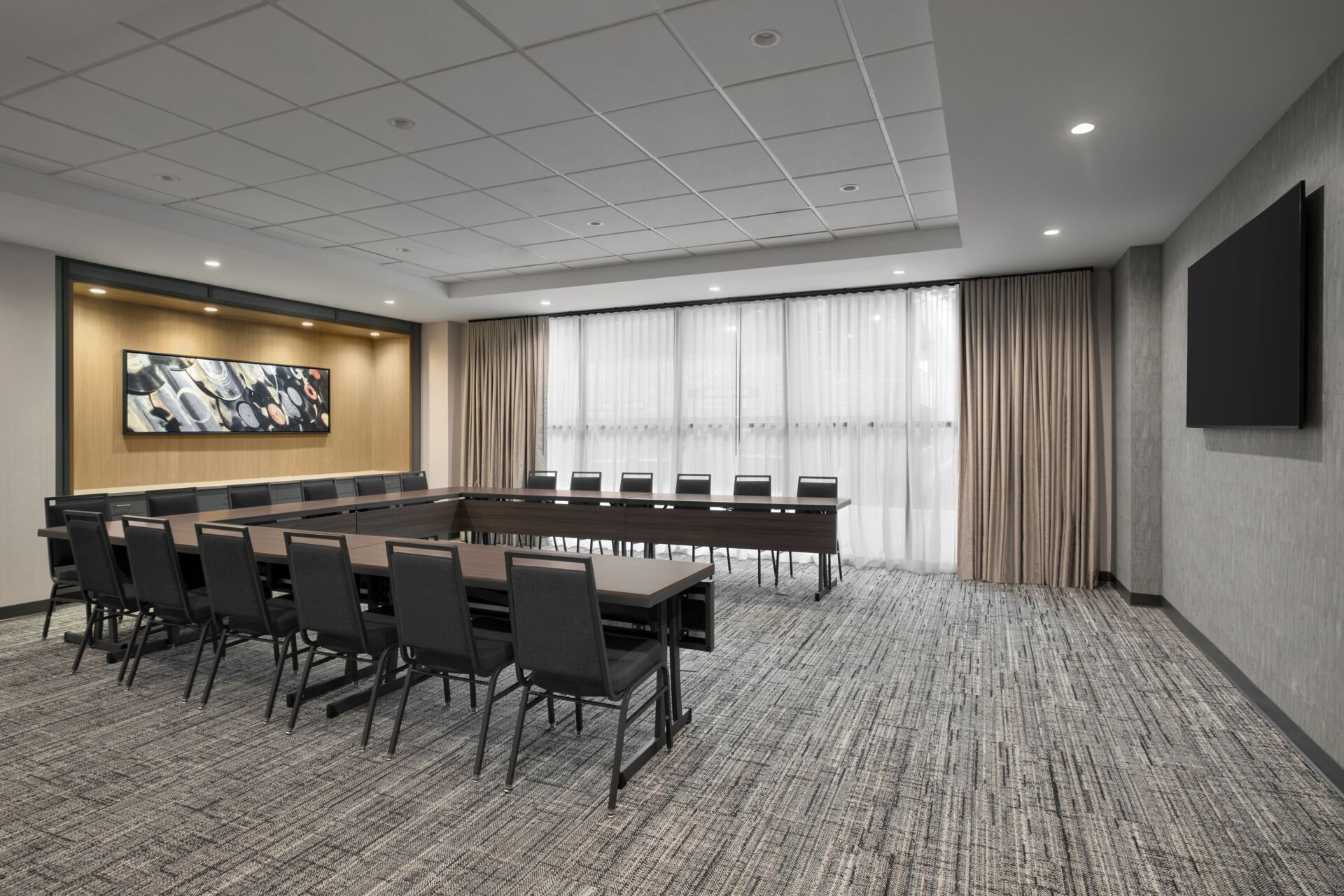 Un salón de conferencias con una larga mesa y sillas, un amplio ventanal con cortinas y una pantalla instalada en la pared.