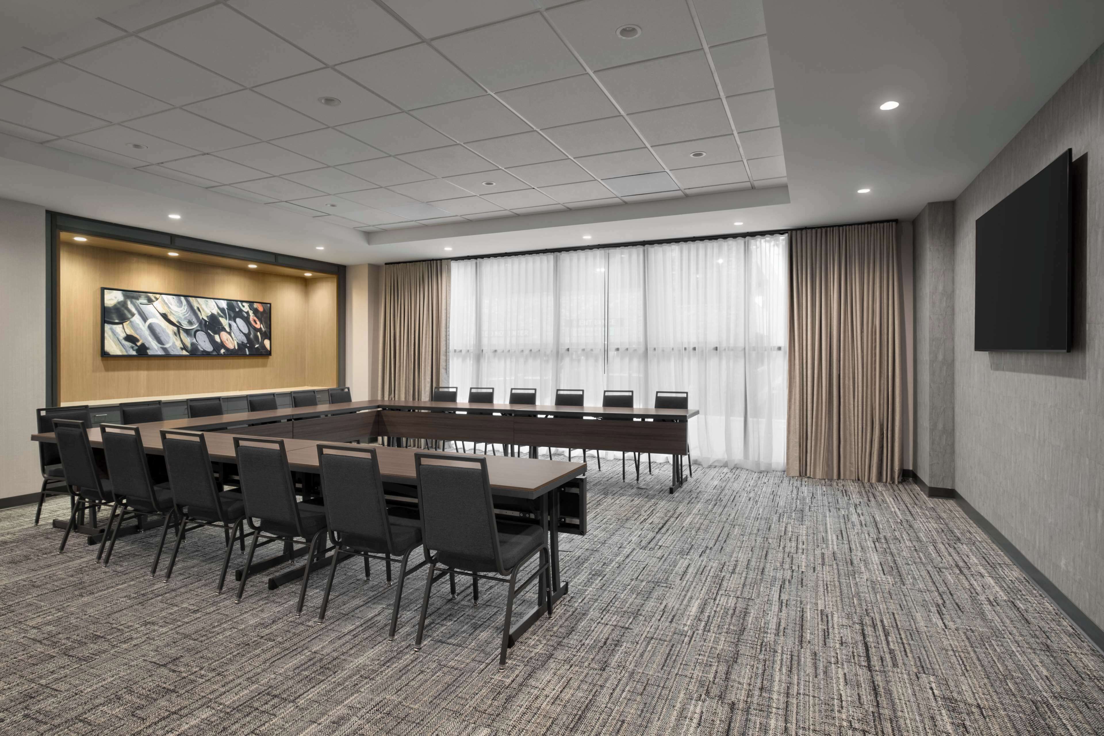 A conference room with a long table, chairs, a large painting, a TV, and a window with curtains.