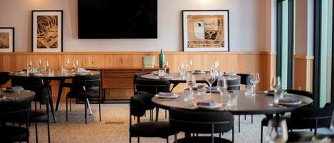 A dining area with round tables, black chairs, and pendant lighting. There are framed artworks on the walls and a large window on the right.