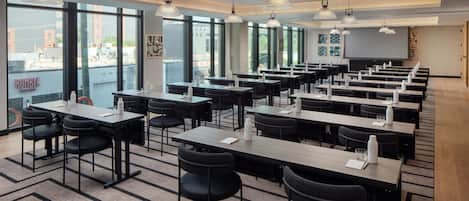 Un salón de conferencias con varias mesas y sillas, ventanas amplias e iluminación colgante.