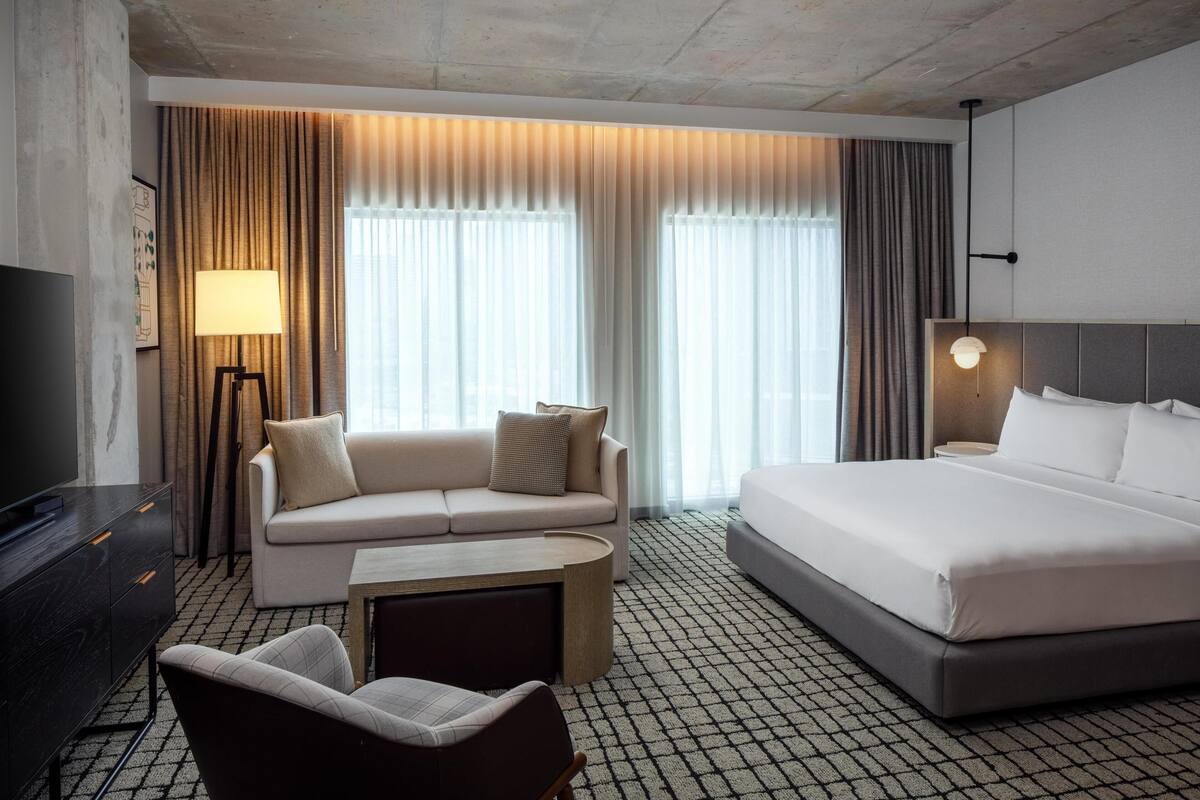 Ein Hotelzimmer mit einem Bett, einer Couch, einem Fernseher, einer Lampe und Vorhängen.