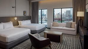Quarto de hotel com duas camas, um sofá, uma poltrona, uma mesinha e janelas amplas com vista para a cidade.