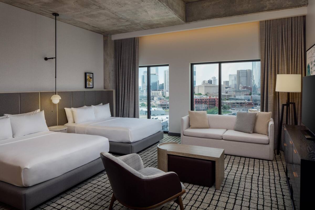 Ein Hotelzimmer mit zwei Betten, einer Couch, einem Sessel, einem kleinen Tisch und großen Fenstern mit Blick auf die Stadt.