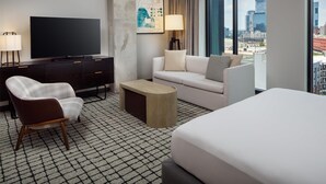 Quarto de hotel moderno com televisão de tela plana grande, sofá, poltrona, mesa de centro e cama.