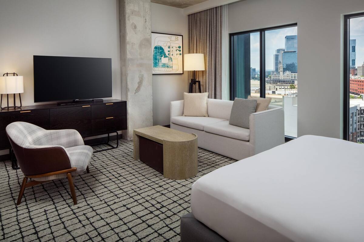 Ein modernes Hotelzimmer mit großem Flachbildfernseher, einer Couch, einem Sessel, einem Couchtisch und einem Bett.