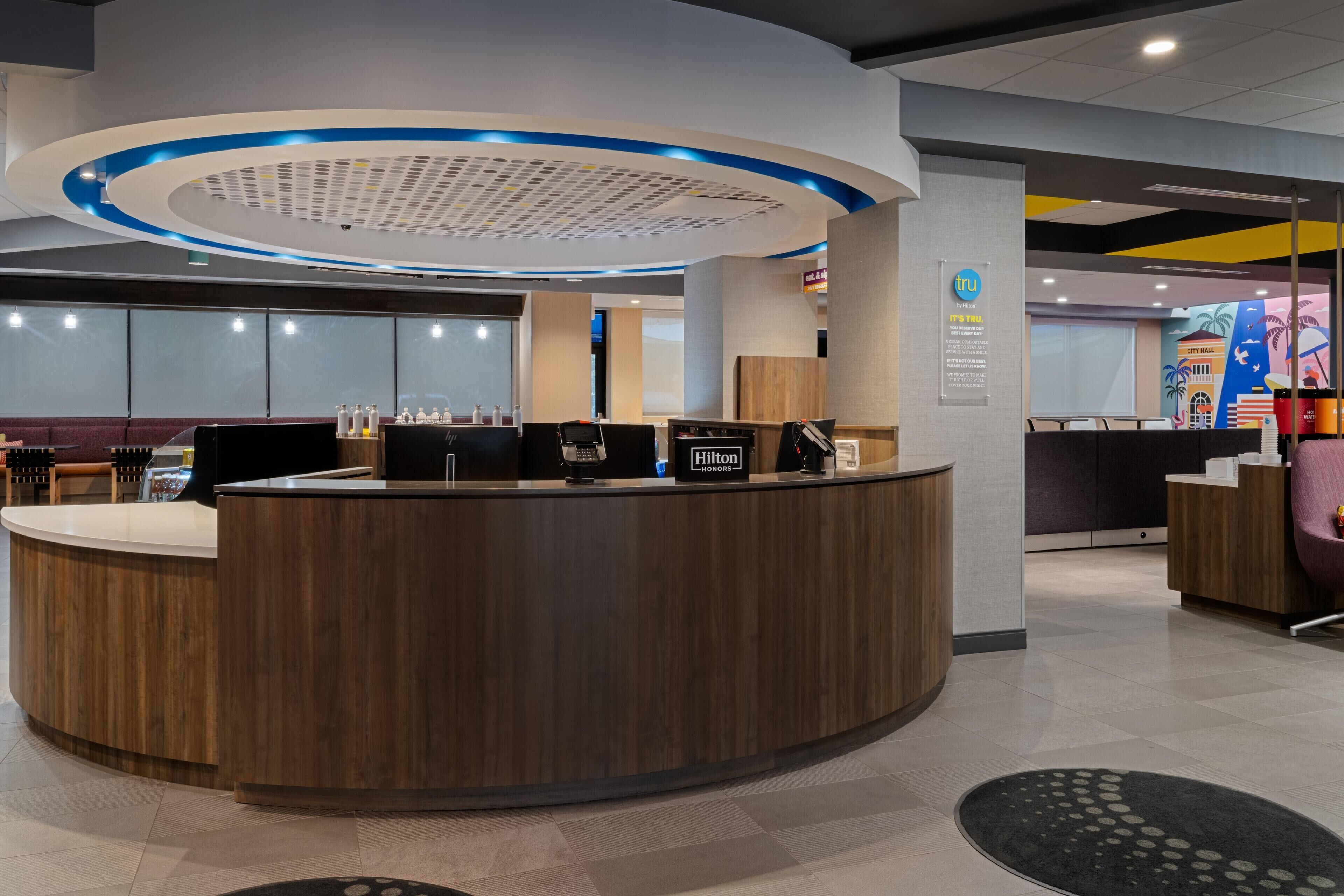 A modern hotel reception area with a wooden reception desk, a digital screen, and a blue-lit ceiling.