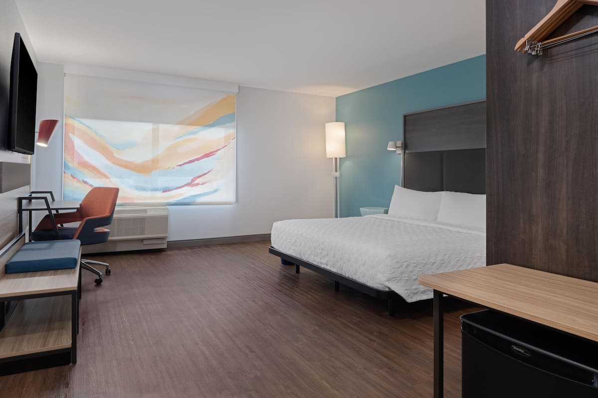 Une chambre d’hôtel moderne, équipée d’un lit, d’un bureau, d’une chaise et ornée d’un grand tableau abstrait au mur.