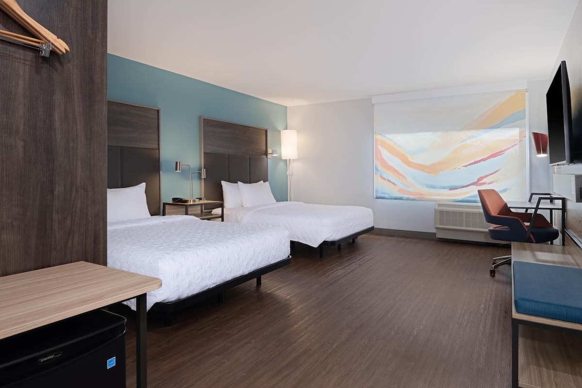 Une chambre d’hôtel avec deux lits, un bureau, une chaise, une télévision et un grand tableau abstrait accroché au mur.