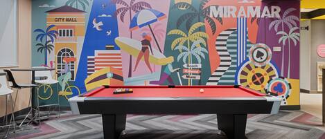 A pool table in a room with a colorful mural depicting a beach scene, including palm trees and a city hall.